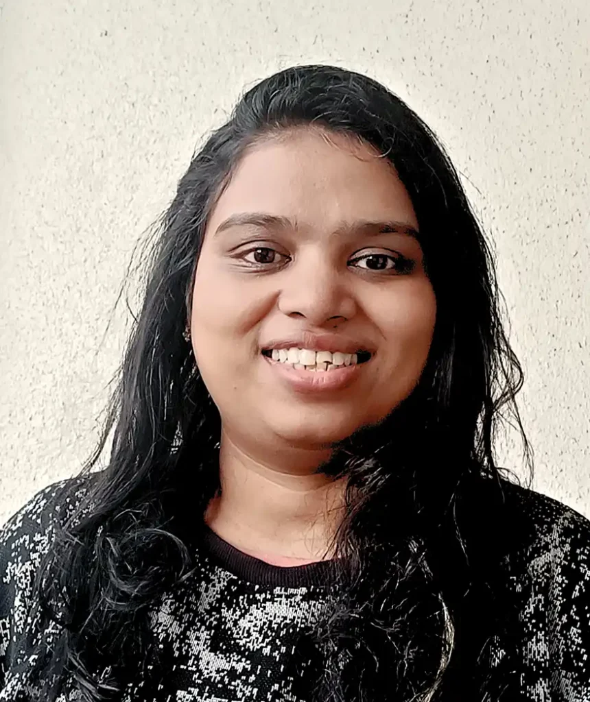Aditi Patil smiling at the camera, wearing a black sweater with a textured pattern, with a light-colored wall in the background.