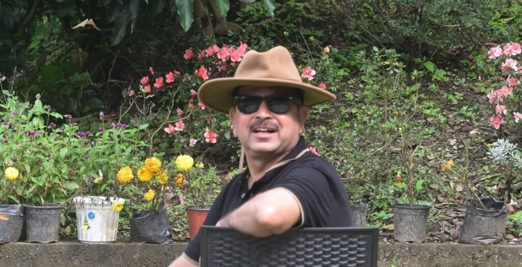A man wearing a brown hat and sunglasses sits outdoors, surrounded by various potted plants and flowers in a garden setting.