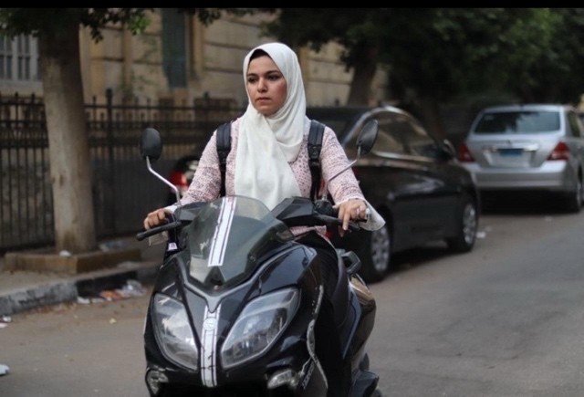 A woman wearing a hijab rides a scooter on a street lined with parked cars, showcasing women's empowerment in urban mobility.