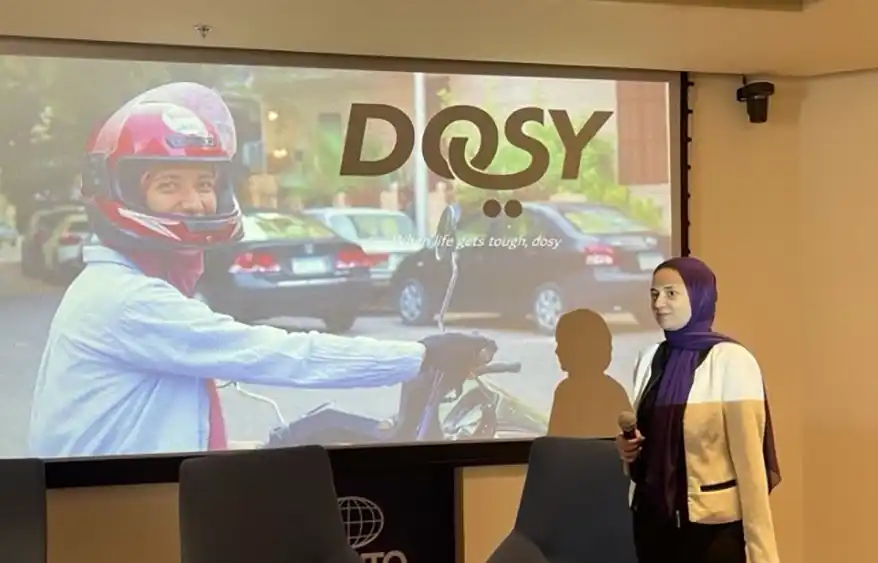 A woman stands in front of a presentation screen displaying the Dosy logo and a smiling rider in a helmet, promoting women's mobility solutions in Egypt.