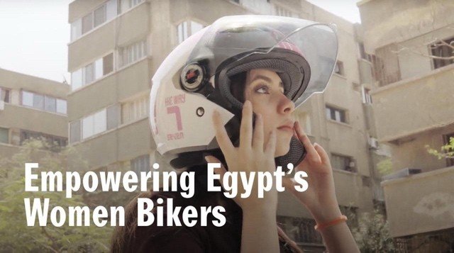 A woman wearing a motorcycle helmet adjusts her headgear, standing in an urban environment with buildings in the background. Text overlay reads 'Empowering Egypt's Women Bikers'.