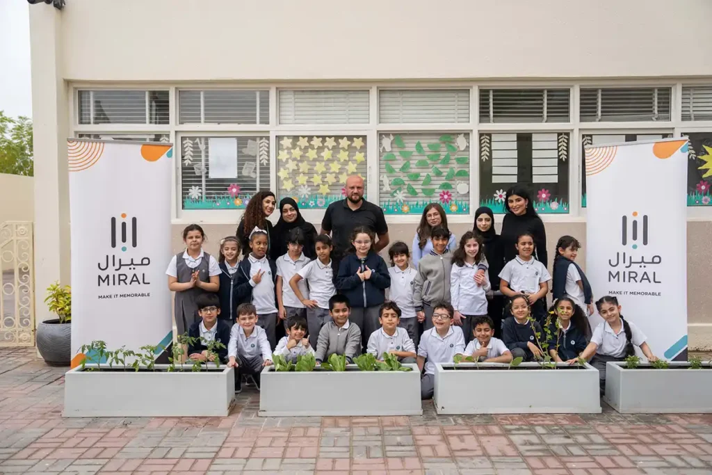 Group of students and educators posing in front of a building, with potted plants and banners for Miral, a community-focused educational initiative in Abu Dhabi.