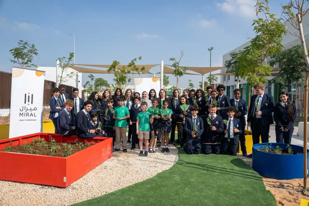 A group of students from various schools in Abu Dhabi, gathered outdoors in a garden setting, participating in the 'Farming in Schools' initiative by Miral. They are holding plants and standing in front of colorful plant beds and a banner showcasing the Miral logo.