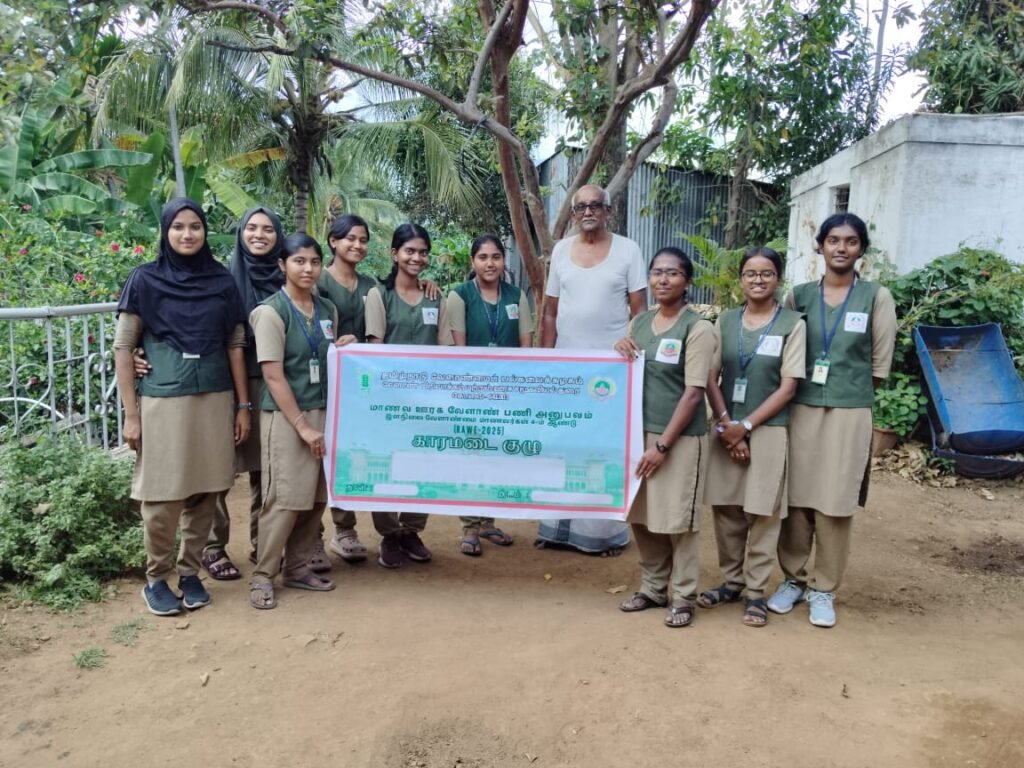 A group of agricultural students standing with a local farmer, holding a banner in Tamil, surrounded by lush greenery and plants.