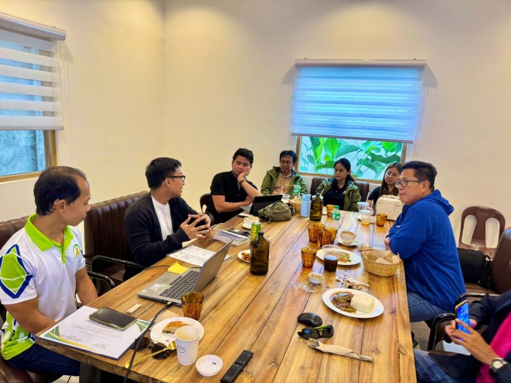 A group of people engaged in a meeting around a wooden table with food and drinks, discussing agricultural and community development initiatives.