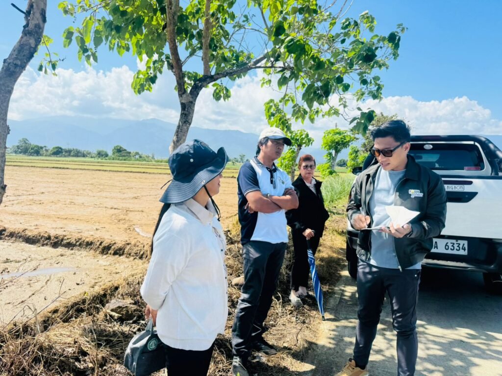 A group of individuals, including a man in a black jacket, discussing plans near a rice field under a blue sky with scattered clouds. Trees are visible in the background.