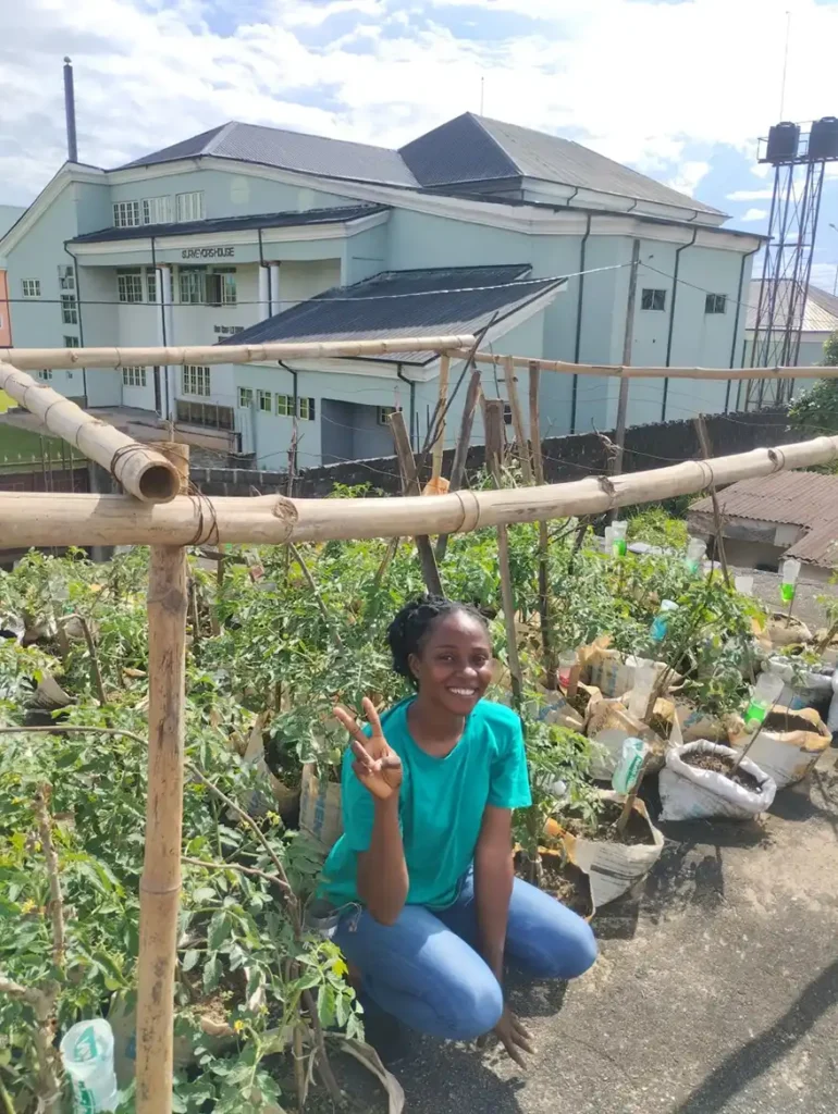 A young woman in a turquoise shirt crouches beside a rooftop garden filled with tomato plants, giving a peace sign. In the background, a building with large windows and a clear blue sky is visible.