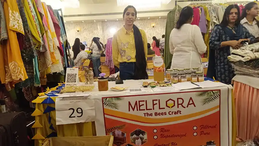 Jasleen standing at the Melflora booth, showcasing raw honey products, at a local exhibition with colorful fabric displays in the background.