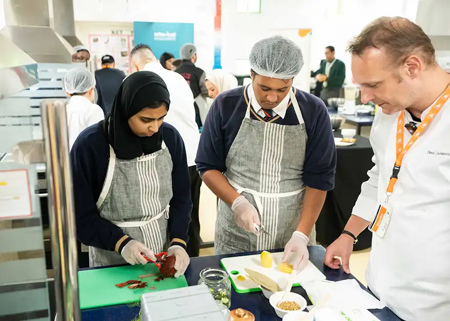 Students participating in a food preservation workshop, preparing ingredients with the guidance of a chef. The setting includes kitchen tools and a focus on sustainable cooking practices.