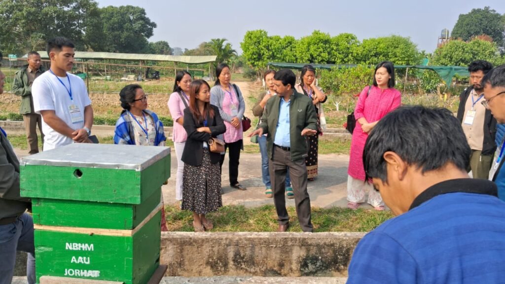 A group of individuals, including men and women, gathered around a green beehive at Assam Agricultural University, guided by a speaker, in an outdoor setting featuring plants and trees.