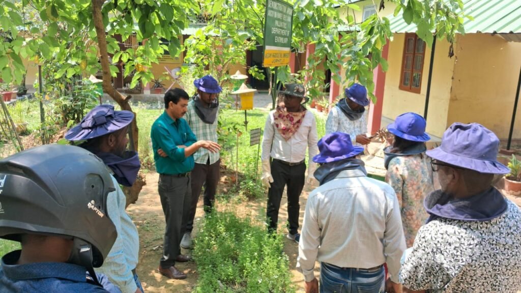 A group of beekeepers in protective gear discussing beekeeping practices in a garden setting, with beekeeping equipment visible in the background.