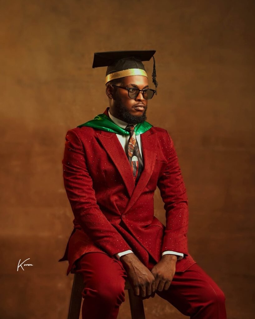 A graduate wearing a red suit and graduation cap, sitting on a stool with a serious expression against a textured background.