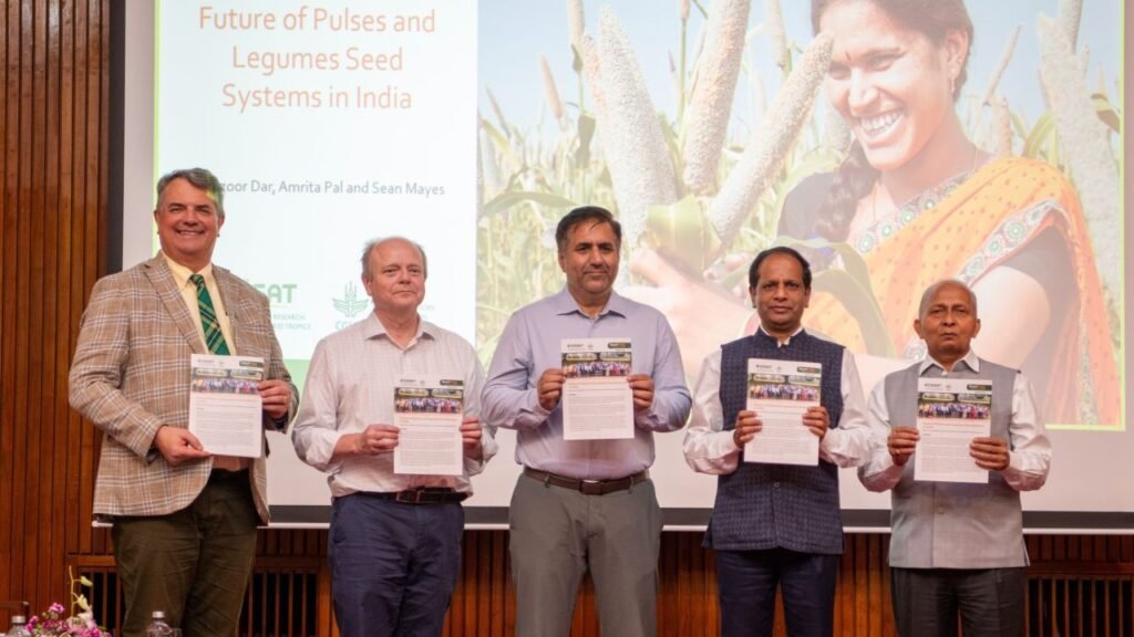 A group of five individuals standing together, holding documents related to agricultural innovations, with a backdrop featuring a smiling woman in a traditional outfit holding crops, conveying themes of collaboration in developing seed systems in India.