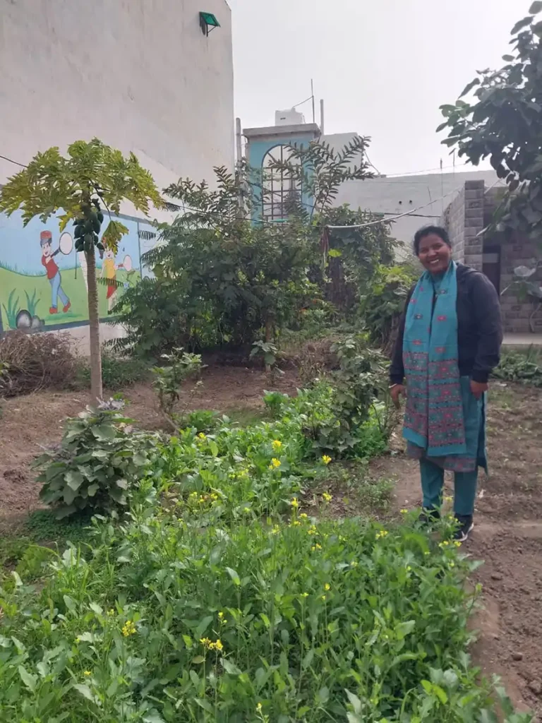 A woman stands in a lush school garden, surrounded by green plants and vegetables, showcasing the initiative of growing food for nutrition in Ghaziabad's government schools.