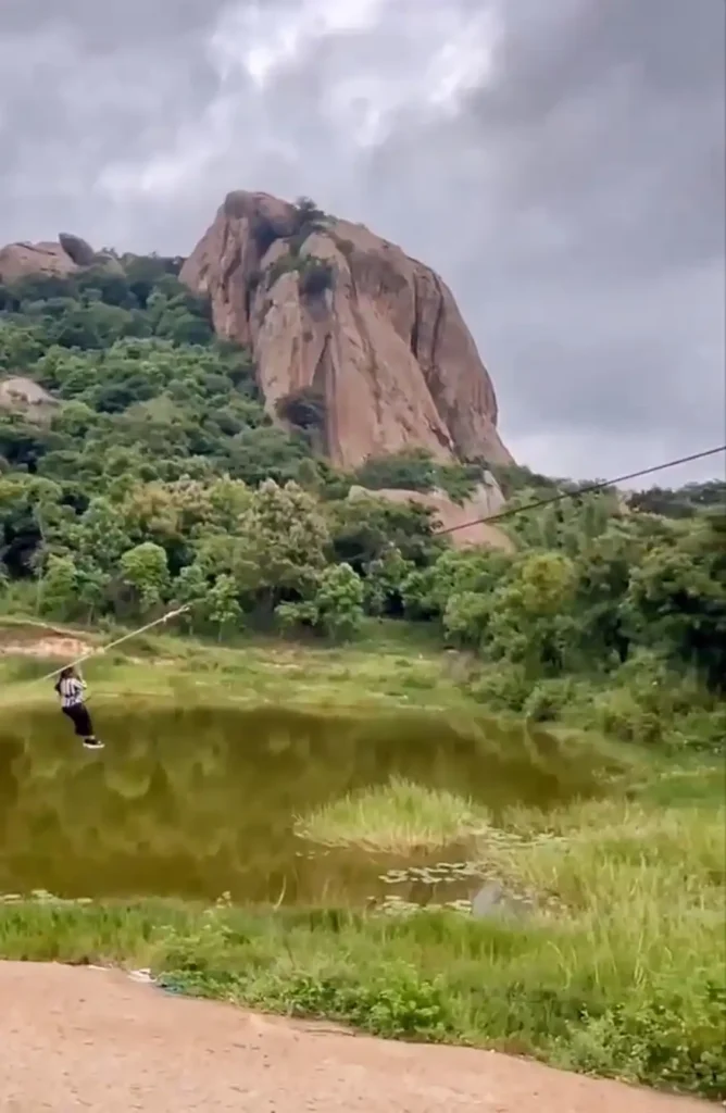 A person zip-lining across a serene landscape in Ramnagar, Karnataka, with rocky cliffs and lush greenery in the background.