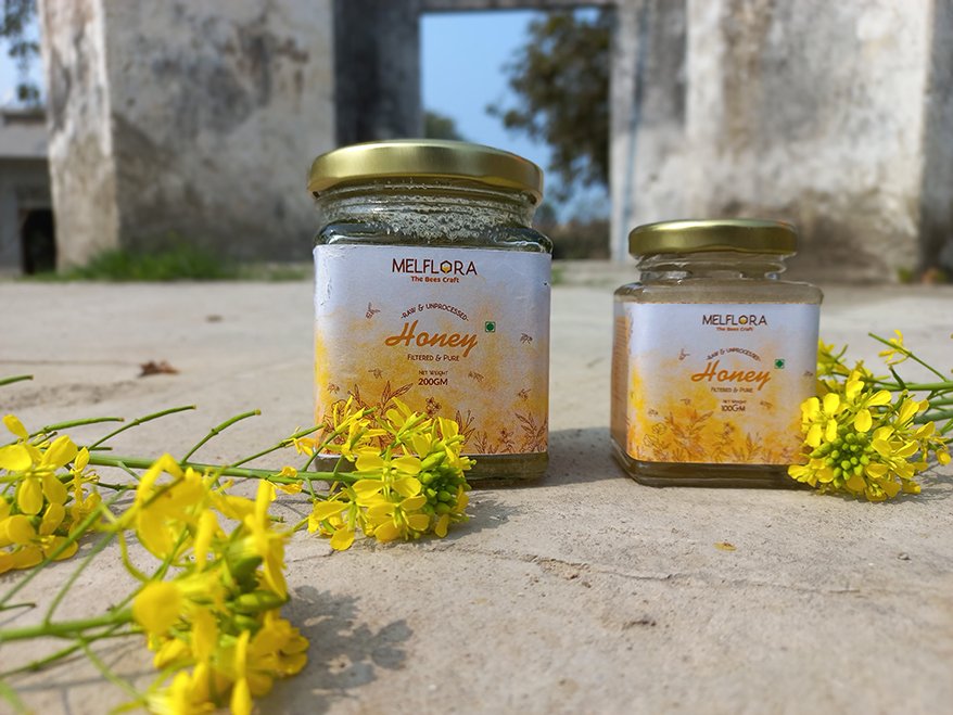 Two jars of Melflora honey labeled 'Raw & Unprocessed Honey' placed on a stone surface with yellow flowers around them.
