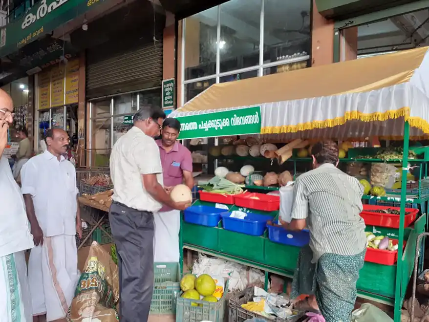 A bustling market scene in Thodupuzha, Kerala, featuring vendors and customers engaging around a green stall displaying a variety of fresh produce, including vegetables and fruits.