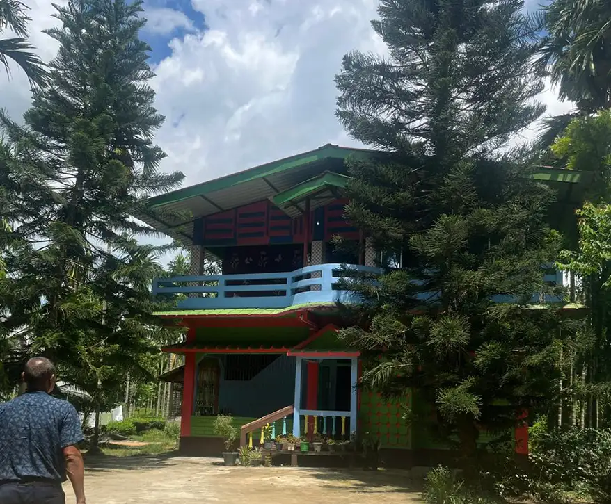 A colorful homestay building surrounded by tall green trees under a blue sky in Barekuri, Assam.