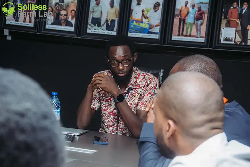 A group discussion at Soilless Farm Lab, featuring a man in a patterned shirt speaking with a serious expression, surrounded by attentive participants. In the background, framed photographs are visible on the wall.