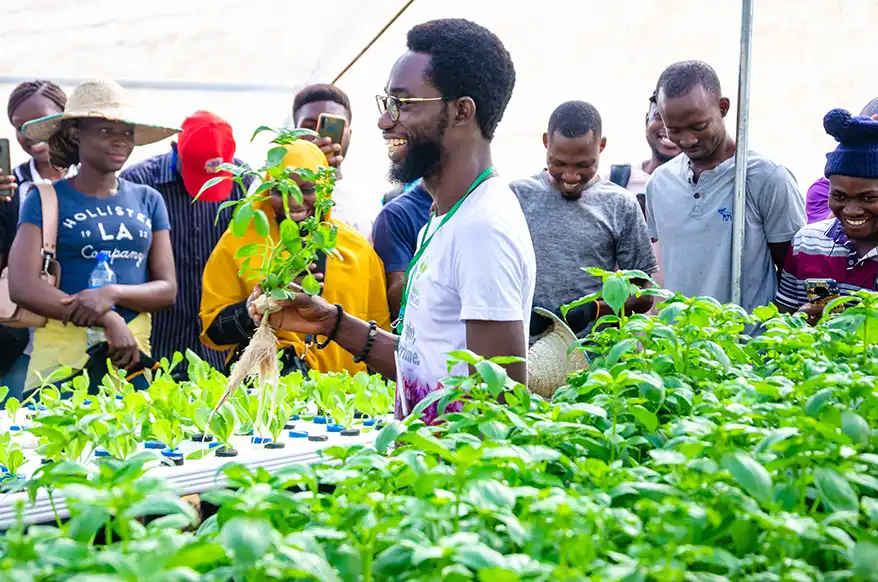 Farmer Samson demonstrating soilless farming techniques to an engaged audience amidst lush green plants in a greenhouse.