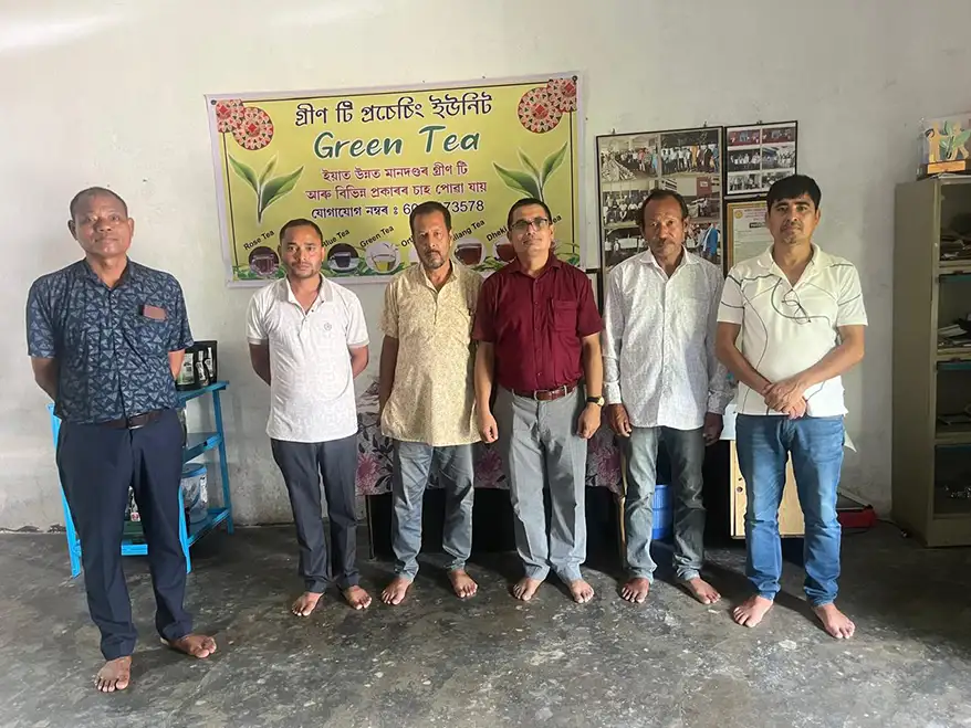A group of six men standing together in an indoor setting, with a sign in the background promoting green tea. The men are dressed in casual attire and barefoot, conveying a relaxed atmosphere.