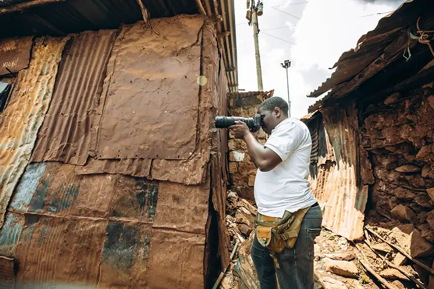 Allan Okumu capturing images in an informal settlement in Nairobi, surrounded by makeshift structures made of metal and wood.