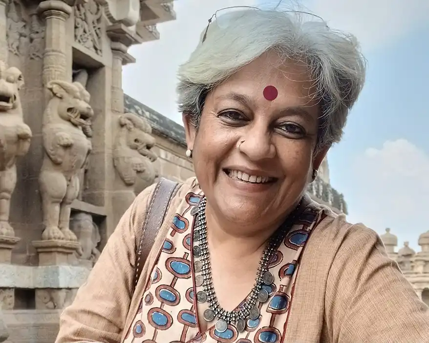 A smiling woman with short grey hair, wearing a traditional outfit adorned with a necklace, poses in front of intricately carved stone structures. She has a decorative red dot on her forehead, symbolizing cultural significance.