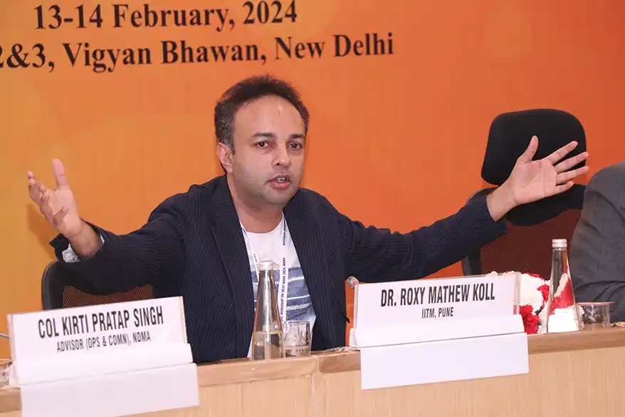 Dr. Roxy Mathew Koll speaking at a conference in New Delhi, gesturing with both hands to emphasize a point while seated at a table with water bottles and papers.
