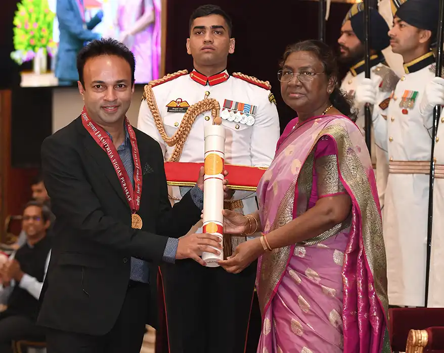 Roxy Mathew Koll receiving an award during a formal ceremony, with a military officer in the background and a woman presenting a scroll.