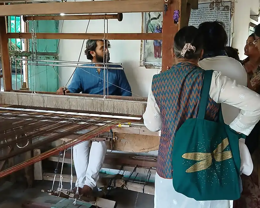 A group of people observing a male artisan at a weaving loom, engaged in traditional textile craft. The artisan is wearing a blue shirt, while the onlookers are in casual clothing, with one carrying a green bag.