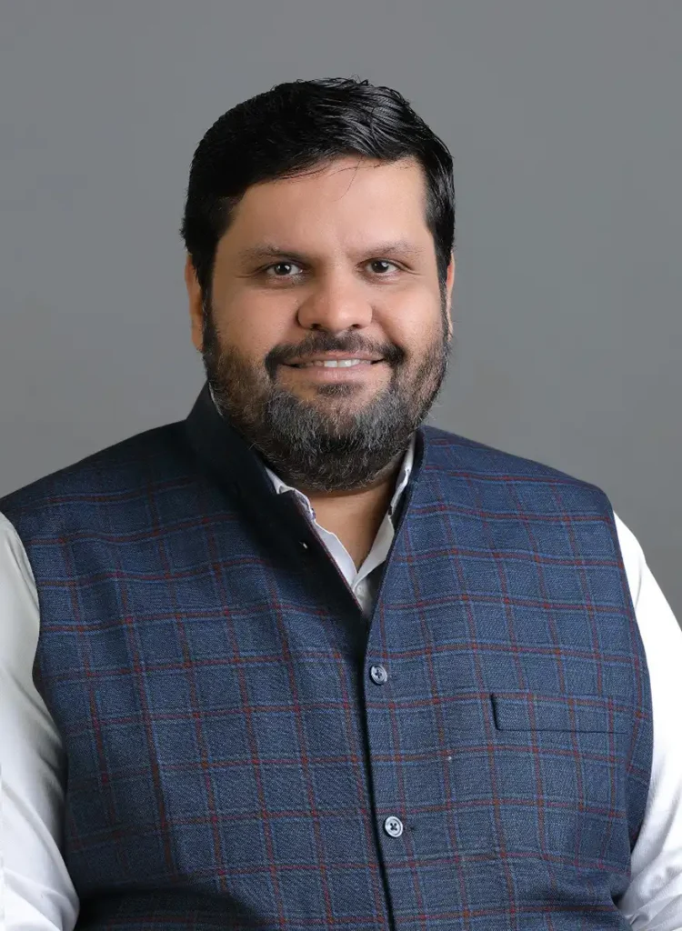 Portrait of Prof. (Dr.) Gourav Vallabh, wearing a dark checkered vest over a white shirt, smiling against a neutral gray background.