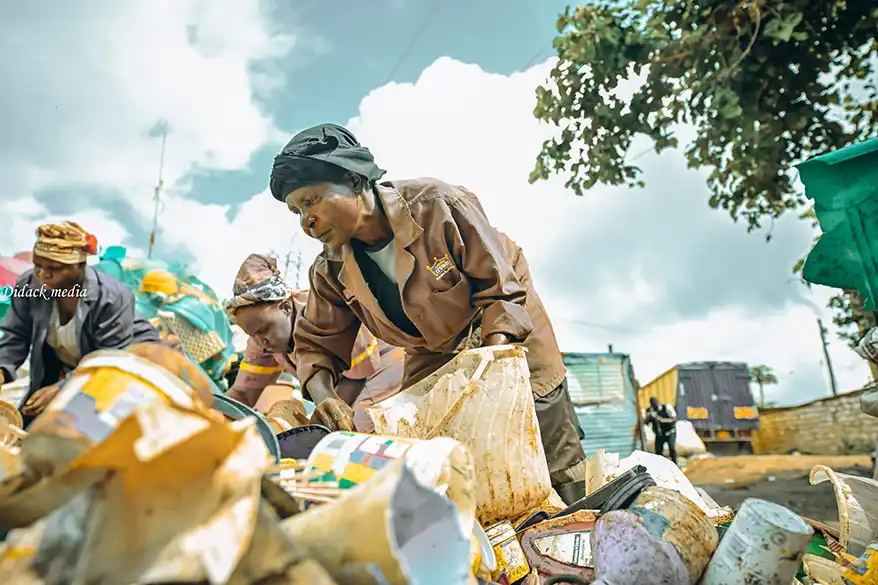 A woman in a brown jacket sorts through a pile of debris and plastic waste in an urban environment, with other workers in the background collecting materials under a partly cloudy sky.
