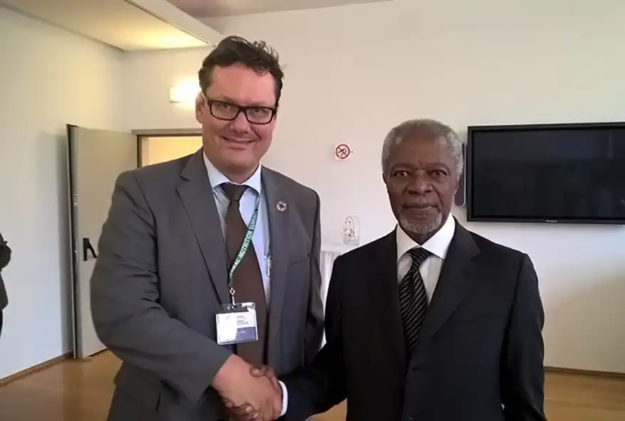 Two men shaking hands in an office setting; one is wearing a suit and tie and the other appears in formal attire.