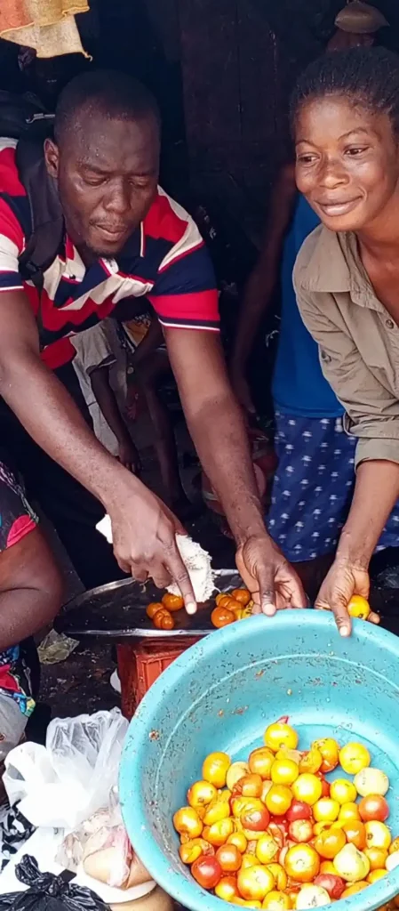 Two traders, a man and a woman, engage in the sale of tomatoes in a bustling market setting, showcasing their goods and interacting with each other. The woman appears to be smiling as she assists in the transaction.