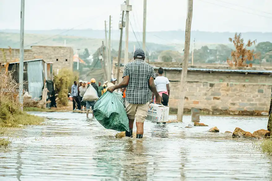 A man walking through floodwaters in Naivasha, carrying a green bag and accompanied by other people navigating a submerged residential area.