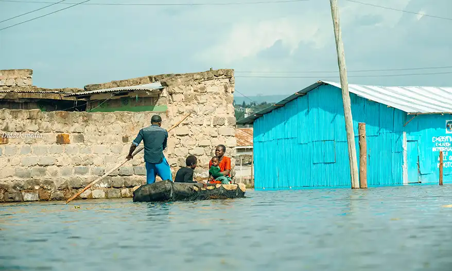 A small boat navigates through flooded residential areas in Naivasha, Kenya, with a man and two children onboard, as surrounding buildings remain partially submerged.