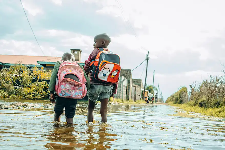 Two children wading through flooded streets, carrying backpacks, with water reaching their calves, amidst a backdrop of homes and power lines.