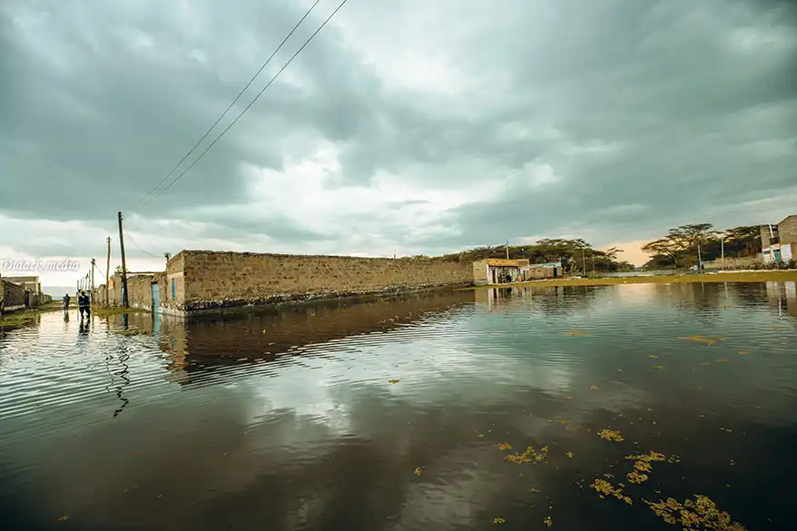 Flooded area in Naivasha, Kenya, with murky water reflecting overcast sky, showing submerged homes and two figures walking through the water.