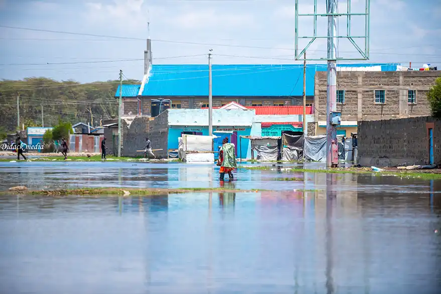 A person walks through a flood-affected area in Naivasha, with buildings partially submerged in water and others visible in the background.