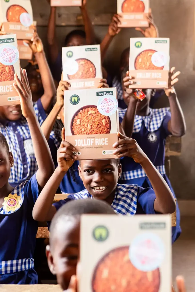 A group of children holding packages of Forti Foods Jollof Rice, smiling and celebrating in a classroom setting.
