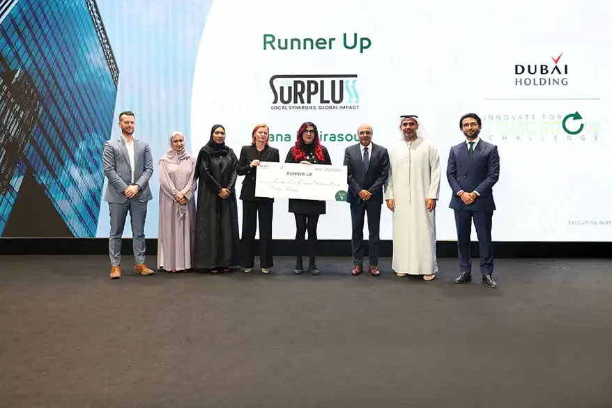 A group of seven individuals stands on stage at the Innovate For Tomorrow Impact Accelerator event, holding a large cheque that reads 'Runner Up' in front of a screen displaying the logos of The Surpluss and Dubai Holding.