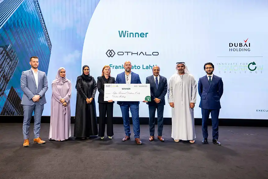 A group of seven people stands on stage at an event, holding a large check made out for AED 500,000. The backdrop displays logos for Othalo and Dubai Holding, with the word 'Winner' prominently visible. The individuals are dressed in formal attire, and the setting appears to be a corporate event focused on innovation and entrepreneurship.