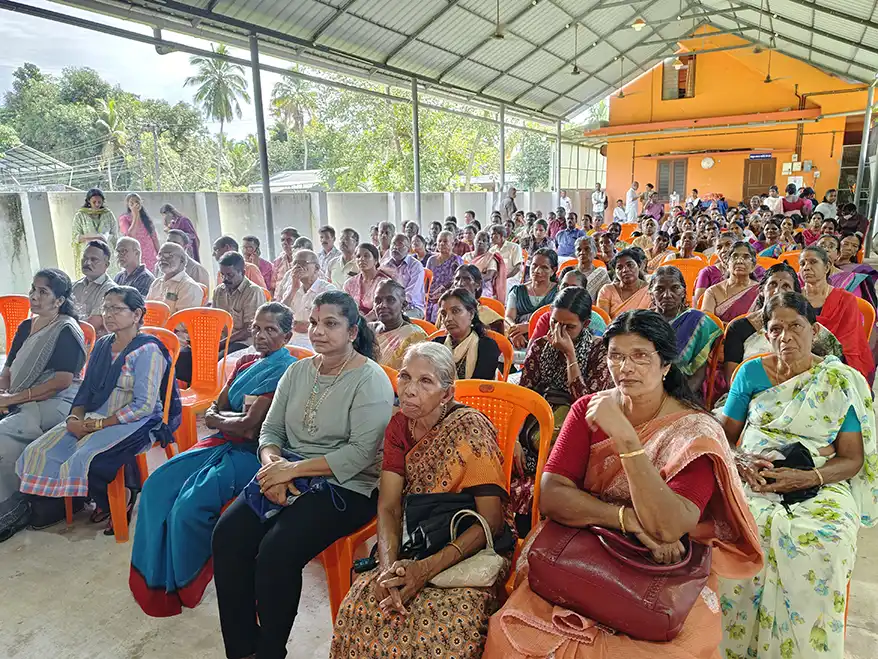 A large gathering of community members seated in an indoor hall, with orange plastic chairs and a bright orange wall in the background. The attendees, primarily women and some men, display a range of expressions, engaged in a meeting or discussion about local issues.