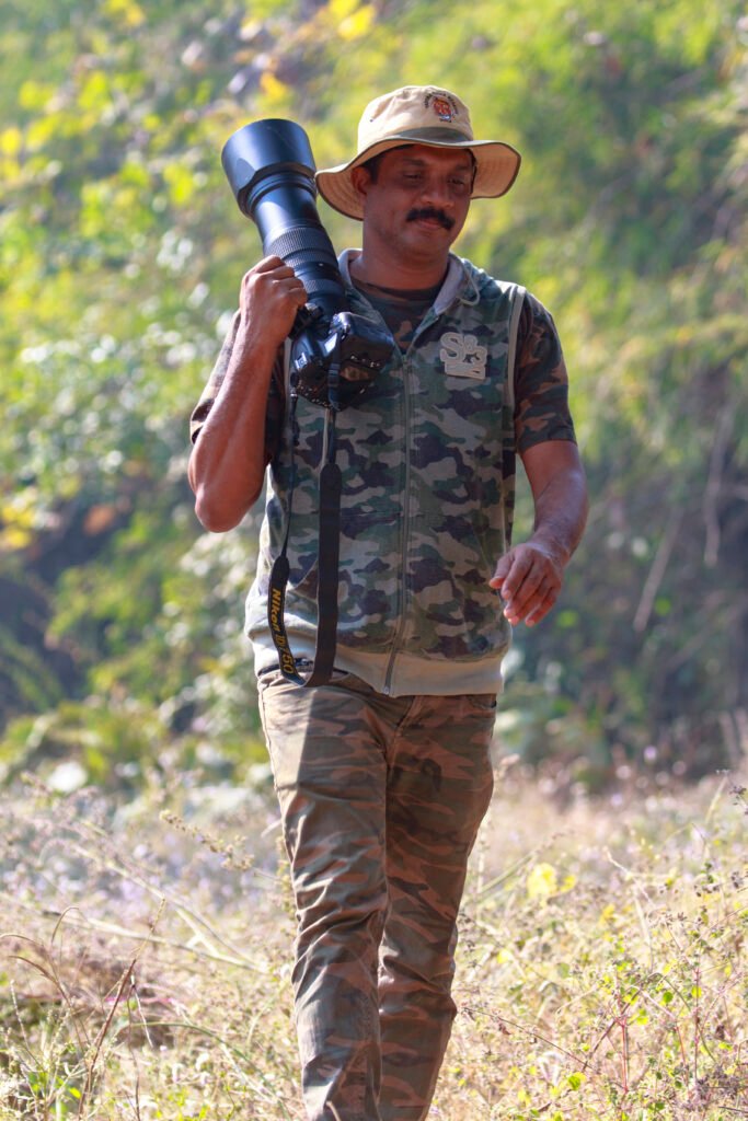 An independent ornithologist and conservationist walking through a forest, carrying a large camera lens.