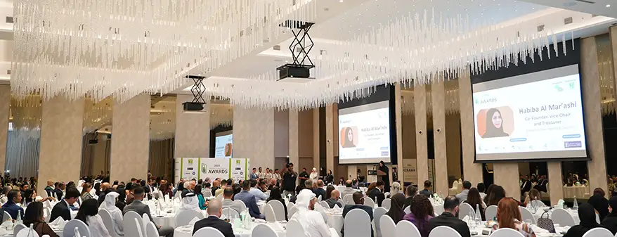 A large audience attending the 2025 MENA Green Building Awards event in Dubai, with a speaker visible on stage and screens displaying information.