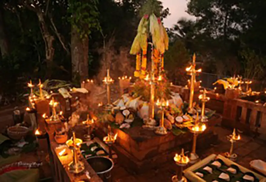 A sacred grove setup during a ceremonial ritual with a decorated serpent idol, surrounded by oil lamps and offerings, in a lush forest setting.