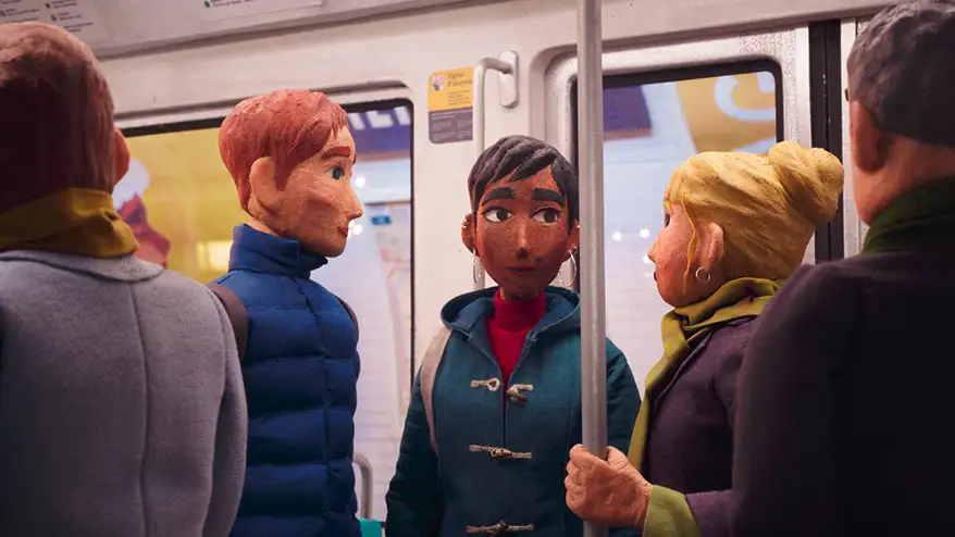 A stop-motion animation scene depicting four characters on a metro, with one woman in the foreground wearing a green coat and looking contemplative, while the others engage in conversation. The setting reflects a multicultural urban environment.