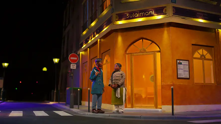 Two animated women stand in front of the 'Sulaimani' restaurant, one dressed in a blue coat and the other in a grey puffer jacket, in a vibrant Parisian street scene at night.