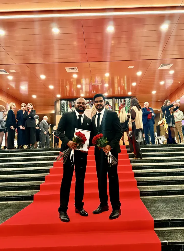 Two men in formal attire stand on a red carpeted staircase, each holding a bouquet of red roses and a certificate. In the background, attendees in elegant outfits are engaged in conversation.