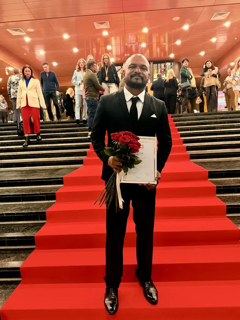 A man in a black suit holds a bouquet of roses and a certificate while standing on red carpet steps. In the background, a crowd of people can be seen at an event.